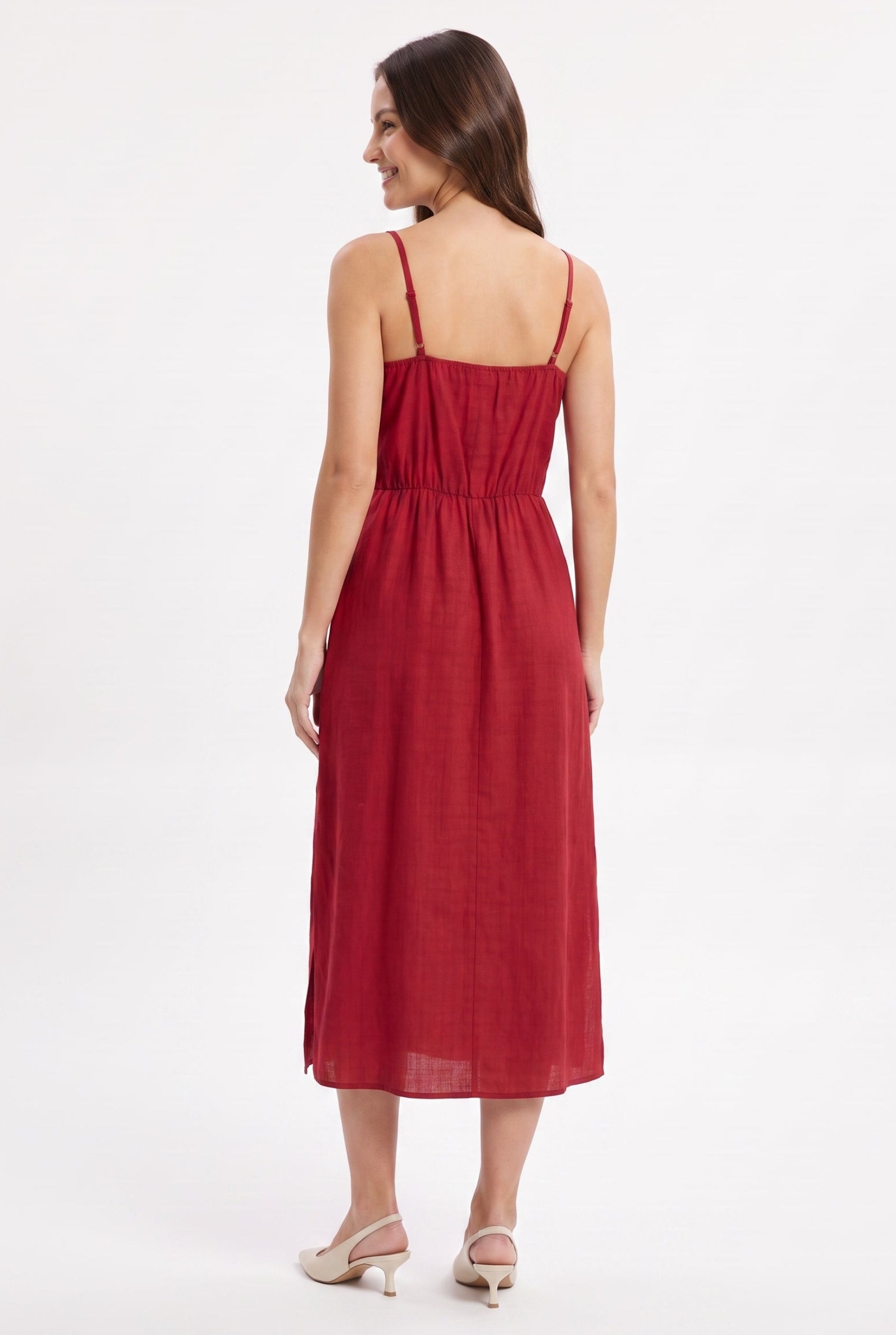 A woman modeling a red midi dress from the back, showing the thin spaghetti straps, gathered waist, and flowing skirt, set against a plain white studio background.